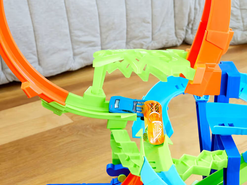 Tor Hot Wheels Stunt Tracks Powietrzna kraksa JFD12 Zbliżenie na część złożonego toru Hot Wheels, gdzie dwa samochodziki (niebieski i pomarańczowy) poruszają się po zielonej i niebieskiej sekcji. W tle widać fragment pętli oraz łóżko/pościel.