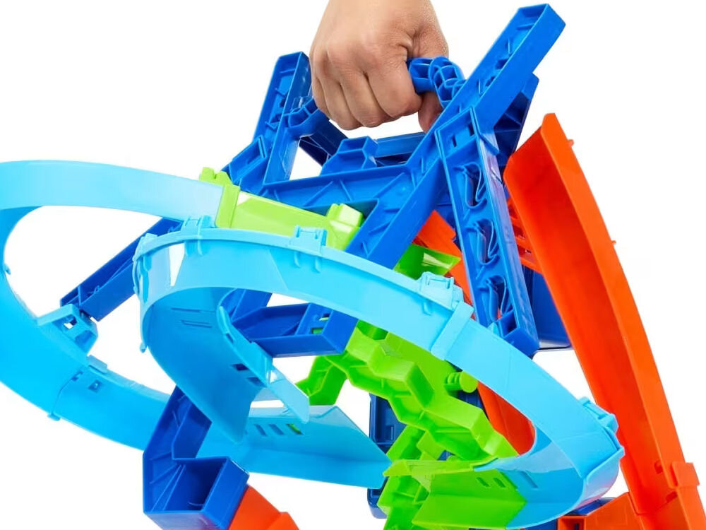 Tor Hot Wheels Stunt Tracks Powietrzna kraksa JFD12 Dłoń trzymająca i podnosząca złożony, wielokolorowy (niebieski, pomarańczowy, zielony) tor wyścigowy Hot Wheels, demonstrując przenośność i kompaktowość zabawki. Tło jest białe.