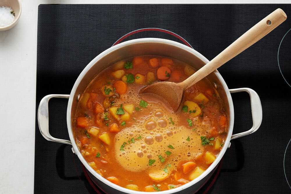 Mata ochronna na płytę indukcyjną LEXPERTO Garnek z gotującą się zupą warzywną ustawiony bezpośrednio na czarnej macie do gotowania na indukcji.