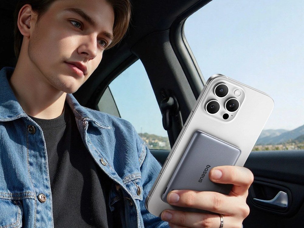 Młody mężczyzna w dżinsowej kurtce trzyma białego iPhone'a z szarym powerbankiem Baseus siedząc w samochodzie.