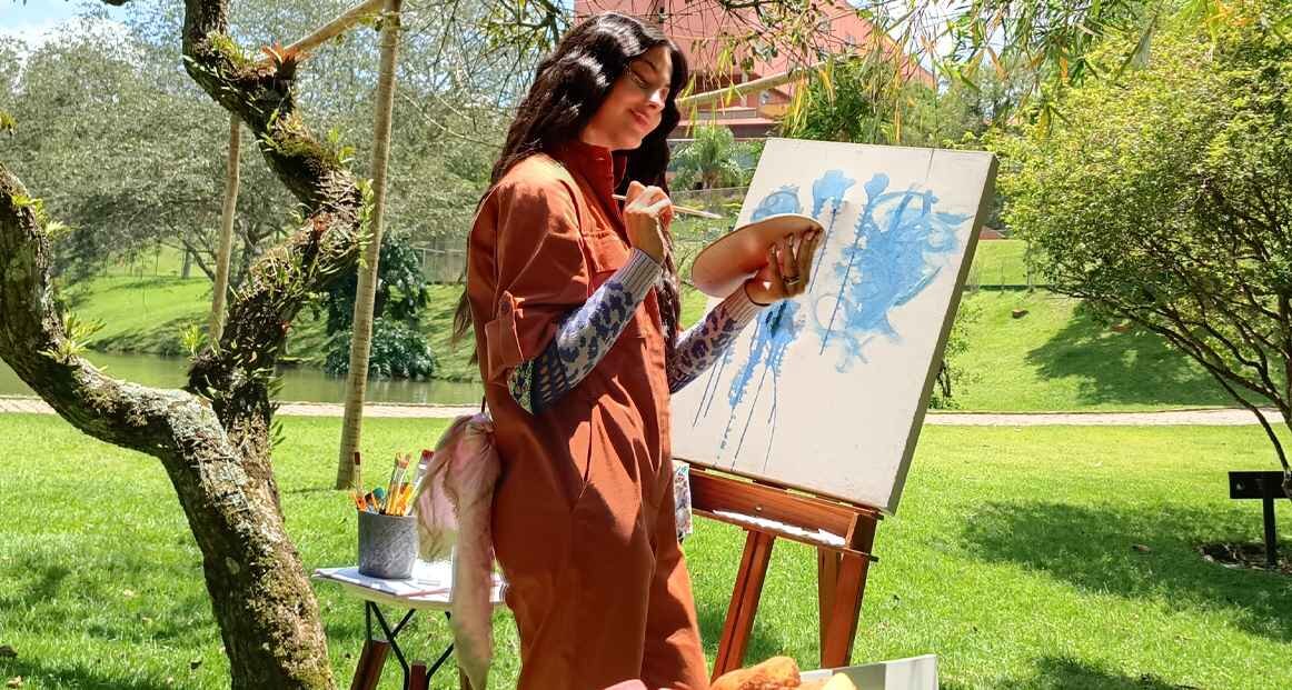 Osoba maluje na płótnie stojącym na sztaludze w plenerze, otoczona zielenią. Na obrazie widoczne są niebieskie abstrakcyjne kształty.