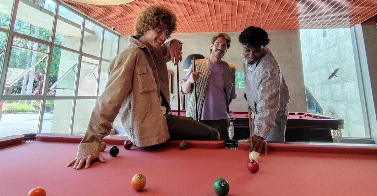 Trzech mężczyzn stoi przy stole bilardowym, jeden z nich trzyma bilę. W tle widać duże okna i ścianę z betonu.