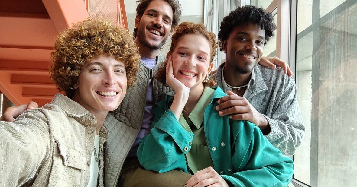 Cztery osoby pozują do zdjęcia w jasnym pomieszczeniu przy oknie. W tle widoczne są pomarańczowe schody i betonowa ściana.