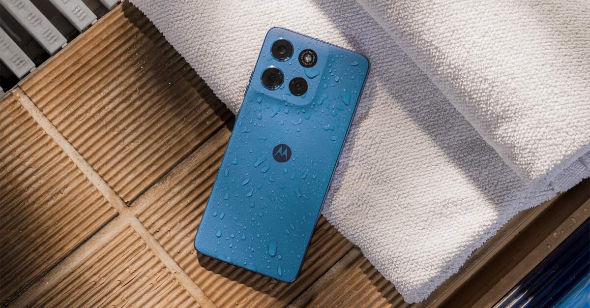 Granatowy smartfon z logo \'M\' na tylnej obudowie, pokryty kroplami wody, leży na białym ręczniku. W tle widoczne są drewniane deski.