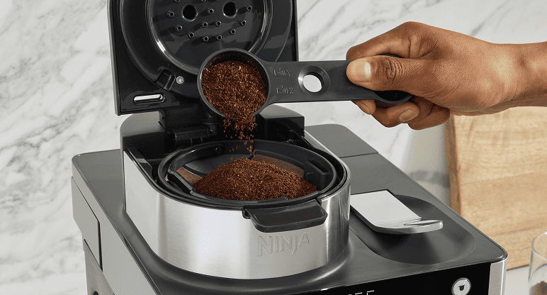 Osoba wsypuje zmieloną kawę za pomocą miarki do koszyka z filtrem w ekspresie Ninja
