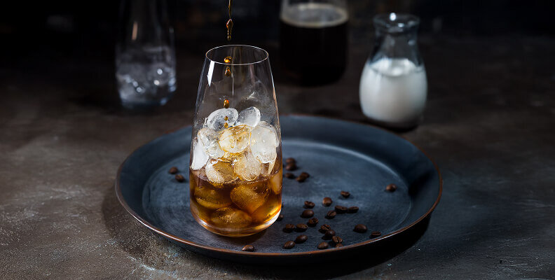 Kawa w stylu cold brew nalewana do wysokiej szklanki wypełnionej lodem, na ciemnej tacy z ziarnami kawy