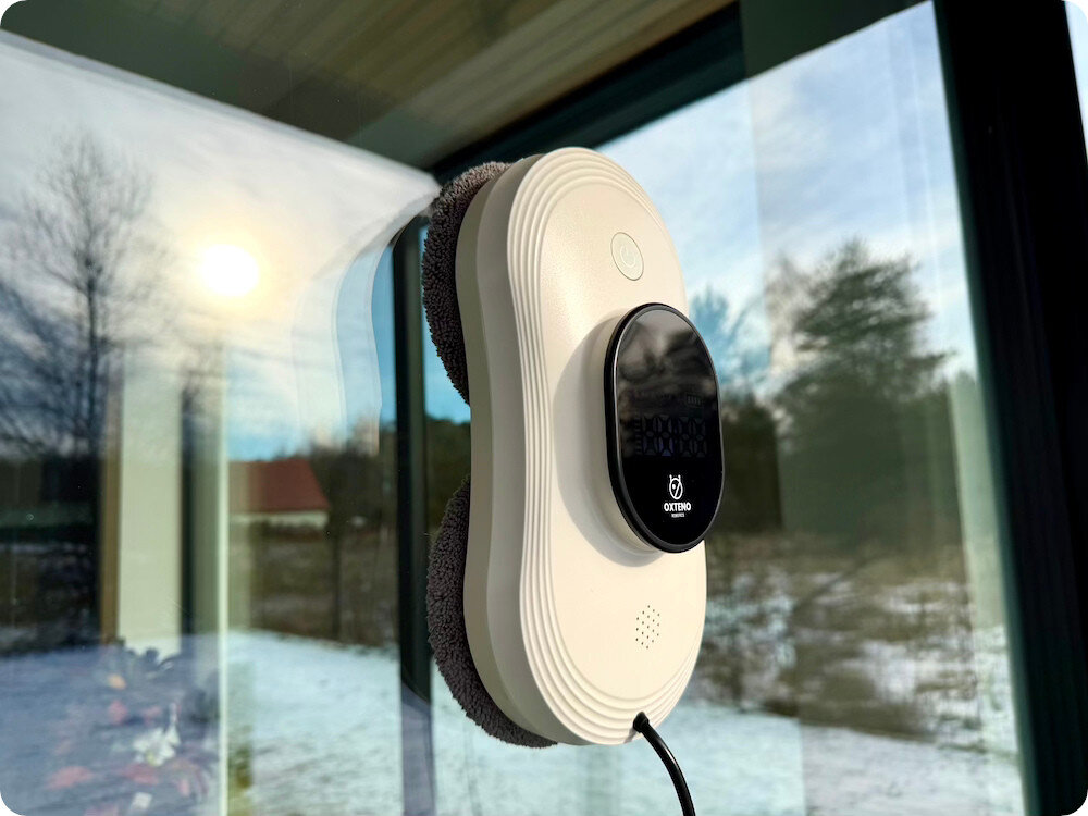 Robot do mycia okien przymocowany do szyby, z widocznym logo OXTENO na czarnej powierzchni. W tle widać odbicie krajobrazu z drzewami i śniegiem.