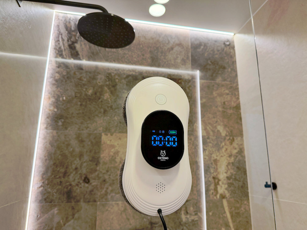 Robot do mycia okien OXTENO OX Glass Pro 3 Elite przymocowany do szklanej powierzchni w łazience. Na wyświetlaczu widoczny jest czas 00:00 oraz logo OXTENO.