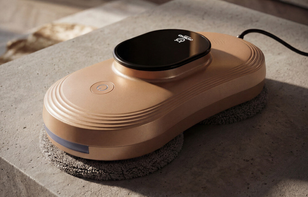 Na betonowej powierzchni znajduje się złoty robot do mycia okien z czarnym panelem na górze, na którym widnieje logo \'OXTENO\'. Urządzenie ma okrągły przycisk i jest podłączone kablem.