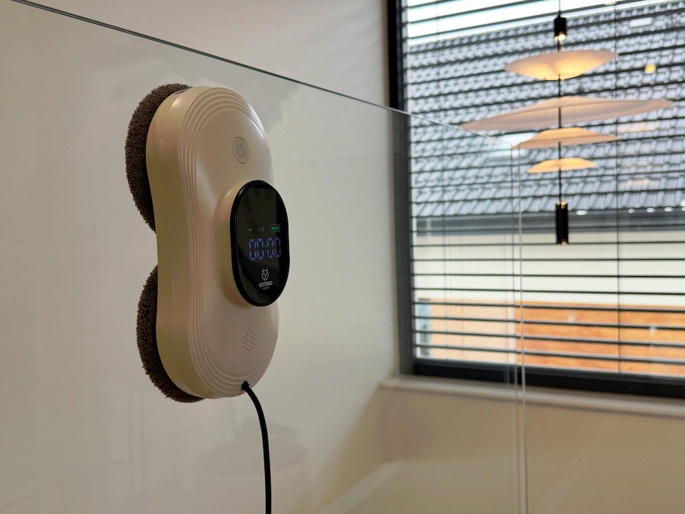 Robot do mycia okien przymocowany do szklanej powierzchni, z widocznym wyświetlaczem i przyciskiem. W tle widać okno z żaluzjami i lampy sufitowe.