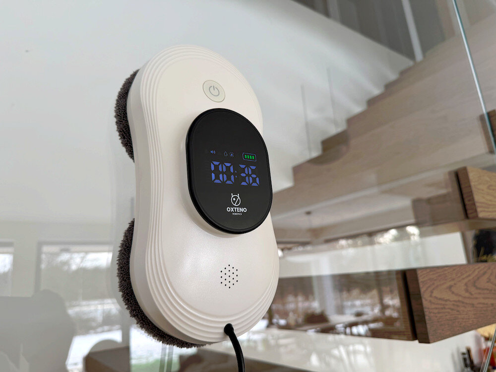 Robot do mycia okien w kolorze białym z wyświetlaczem pokazującym czas 00:36, zamocowany na szklanej powierzchni. Na wyświetlaczu widoczne jest logo OXTENO.