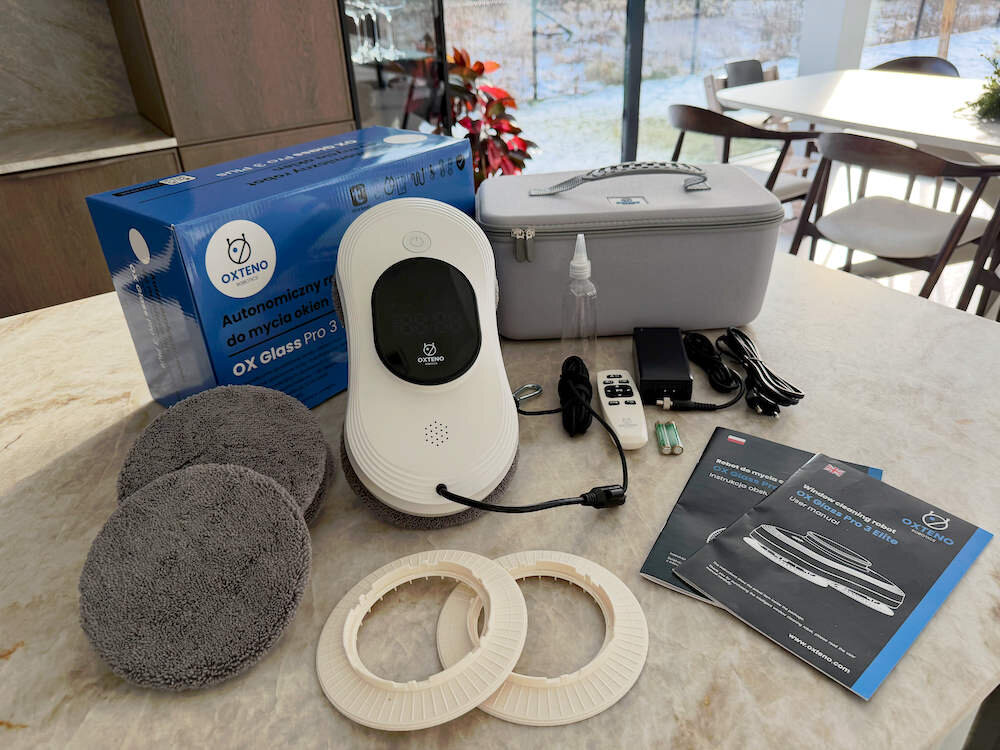 Na stole znajduje się zestaw do mycia okien, w tym robot OXTENO, szare ściereczki, pierścienie, pilot, ładowarka, butelka z rozpylaczem oraz instrukcje obsługi. W tle widoczna jest niebieska skrzynka z napisem \'OXTENO Automatyczny robot do mycia okien OX Glass Pro 3 Elite\'.