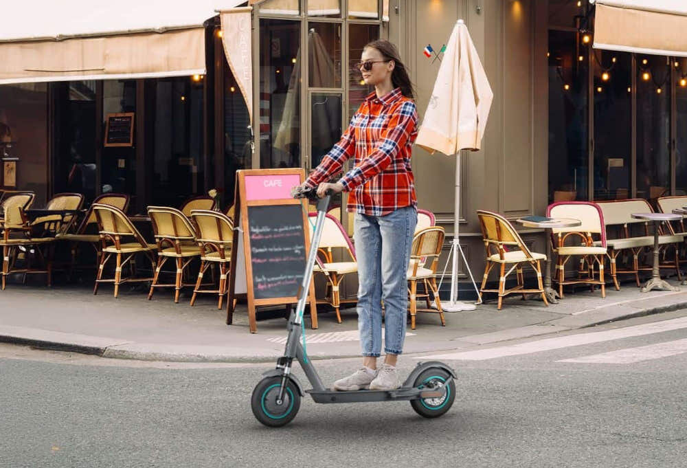 Hulajnoga elektryczna MOTUS Scooty 10 Gen 6 65km 350W 10 cali Szary miasto kobieta jedzie na hulajnodze w tle restauracja 3 tryby jazdy przycisk POWER umieszczonym na wyświetlaczu tryb pieszy prędkość do 6 km/h tryb standardowy prędkość do 15 km/h tryb sportowy prędkość do 20 km/h