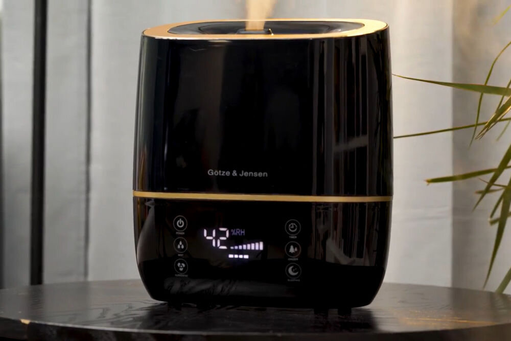 Nawilżacz GÖTZE & JENSEN AH250K Zbliżenie na czarny, błyszczący nawilżacz powietrza Gotze & Jensen ze złotym wykończeniem. Wyświetlacz pokazuje 42% wilgotności względnej (RH) i smugę pary wodnej unoszącą się z góry urządzenia.