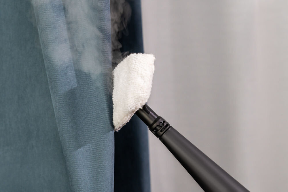 ODKURZACZ PIORĄCY SPOTCLEAN GOTZE&JENSEN CSC600 z funkcją czyszczenia parowego do odświeżania tkanin firanek zasłon eliminowania bakterii roztoczy alergenów Zbliżenie na czarną końcówkę parową z nałożoną białą nakładką z mikrofibry, która jest używana do odświeżania niebieskiej zasłony (pary z urządzenia są widoczne).