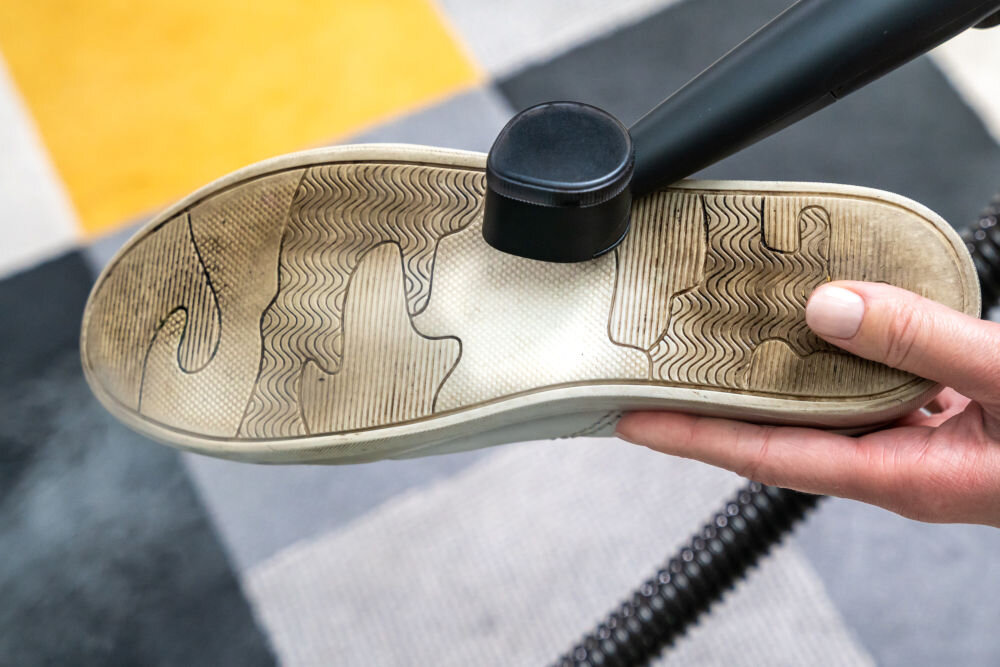 ODKURZACZ PIORĄCY SPOTCLEAN GOTZE&JENSEN CSC600 ze szczotką uniwersalną szeroką do butów do czyszczenia tapicerki meblowej samochodowej dywanów Zbliżenie na dłoń trzymającą podeszwę brudnego, białego buta, którą jest czyszczona małą czarną końcówką odkurzacza.