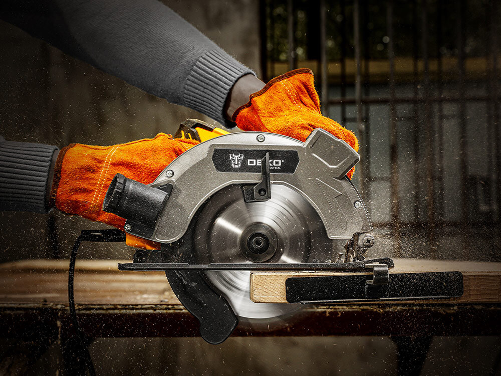 Pilarka tarczowa DEKO DKCS16G185, pilarka, tarcza, cięcie, Pilarka tnie drewo w rękach osoby z założonymi rękawicami ochronnymi, w tle plac budowy