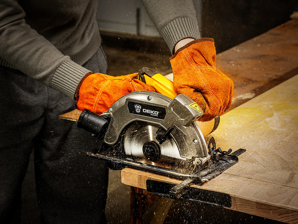 Pilarka tarczowa DEKO DKCS16G185, wszechstronność, silnik, prędkość, cięcie, komfort, ergonomia, Osoba obsługuje pilarkę która tnie deskę na stole roboczym, w tle plac budowy