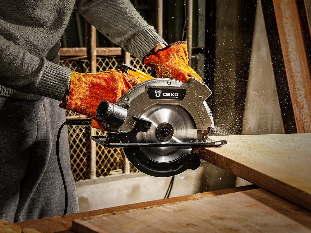 Pilarka tarczowa DEKO DKCS16G185, przycisk, bezpieczeństwo, uchwyty, komfort, Pilarka w rękach osoby, rozpoczyna tnięcie drewnna na blacie, w tle plac budowy