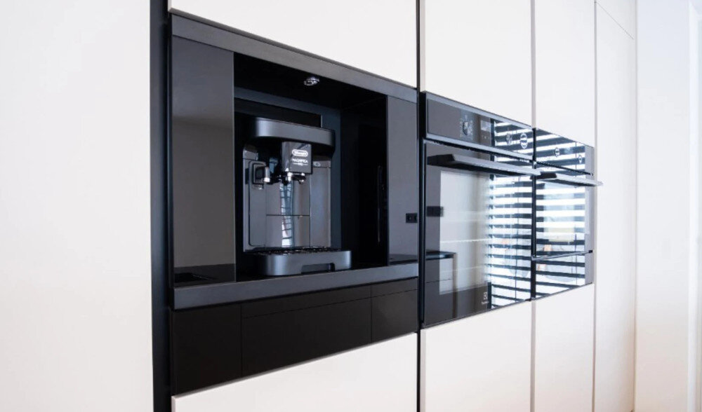 Szuflada do ekspresu KLUGE KFB01BB Na zdjęciu przedstawiono elegancką zabudowę kuchenną z jasnymi frontami, w której centralnie umieszczony jest czarny ekspres do kawy w niszy. Obok znajdują się dwa piekarniki lub piekarnik z kuchenką mikrofalową w czarnym szkle, tworząc spójną, minimalistyczną linię sprzętów dla miłośników kawy dodatek do ekspresu wolnostojącego aranżacja kuchenna