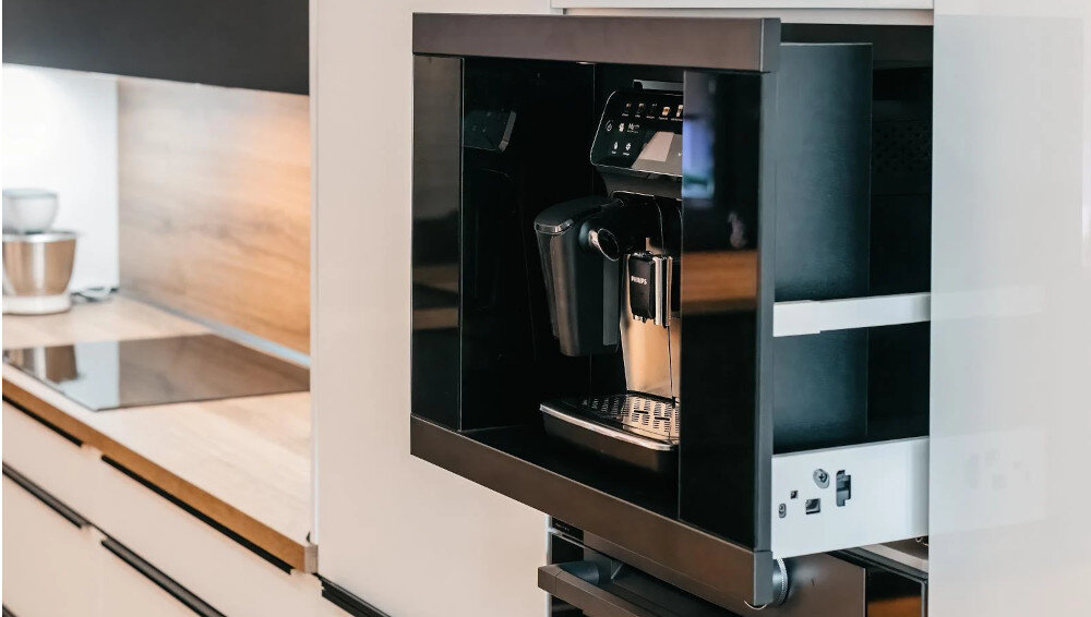 Szuflada do ekspresu KLUGE KFB01BB Na zdjęciu widoczny jest czarny ekspres do kawy wysunięty na prowadnicach z zabudowy meblowej, co ułatwia dostęp do urządzenia. W tle widać kuchnię z drewnianym blatem, płytą grzewczą i jasnymi frontami szafek praktyczny stylowy dodatek trwałe prowadnice teleskopowe 