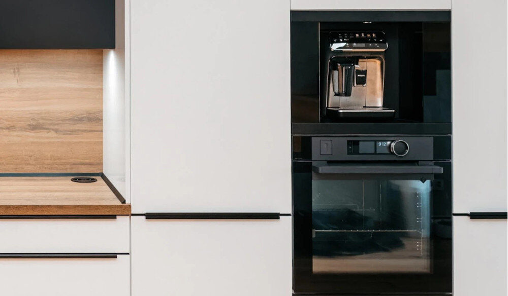 Szuflada do ekspresu KLUGE KFB03BB Na zdjęciu widać zabudowę kuchenną z białymi frontami i drewnianym blatem, podświetloną listwą LED nad przestrzenią roboczą. W centralnej części słupa meblowego umieszczono czarny piekarnik do zabudowy, a nad nim ekspres do kawy, tworząc spójną, elegancką linię sprzętów