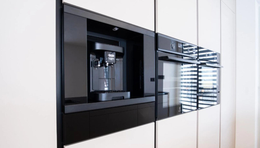Szuflada do ekspresu KLUGE KFB03BB Na zdjęciu przedstawiono elegancką zabudowę kuchenną z jasnymi frontami, w której centralnie umieszczony jest czarny ekspres do kawy w niszy. Obok znajdują się dwa piekarniki lub piekarnik z kuchenką mikrofalową w czarnym szkle, tworząc spójną, minimalistyczną linię sprzętów dla miłośników kawy dodatek do ekspresu wolnostojącego aranżacja kuchenna