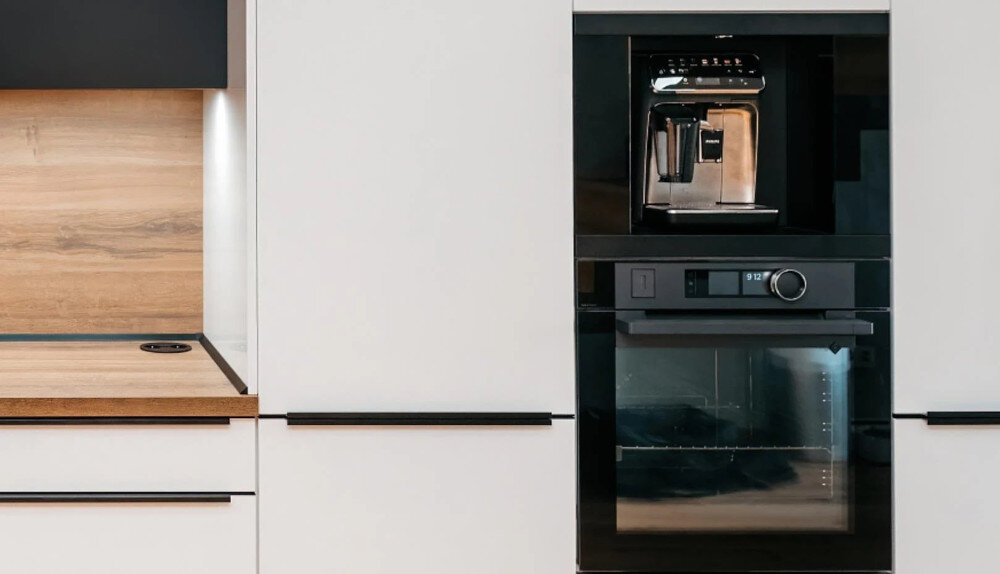Szuflada do ekspresu KLUGE KFB07BB Na zdjęciu widać zabudowę kuchenną z białymi frontami i drewnianym blatem, podświetloną listwą LED nad przestrzenią roboczą. W centralnej części słupa meblowego umieszczono czarny piekarnik do zabudowy, a nad nim ekspres do kawy, tworząc spójną, elegancką linię sprzętów