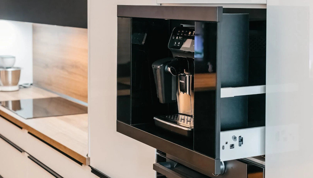 Szuflada do ekspresu KLUGE KFB07BB Na zdjęciu widoczny jest czarny ekspres do kawy wysunięty na prowadnicach z zabudowy meblowej, co ułatwia dostęp do urządzenia. W tle widać kuchnię z drewnianym blatem, płytą grzewczą i jasnymi frontami szafek praktyczny stylowy dodatek trwałe prowadnice teleskopowe 