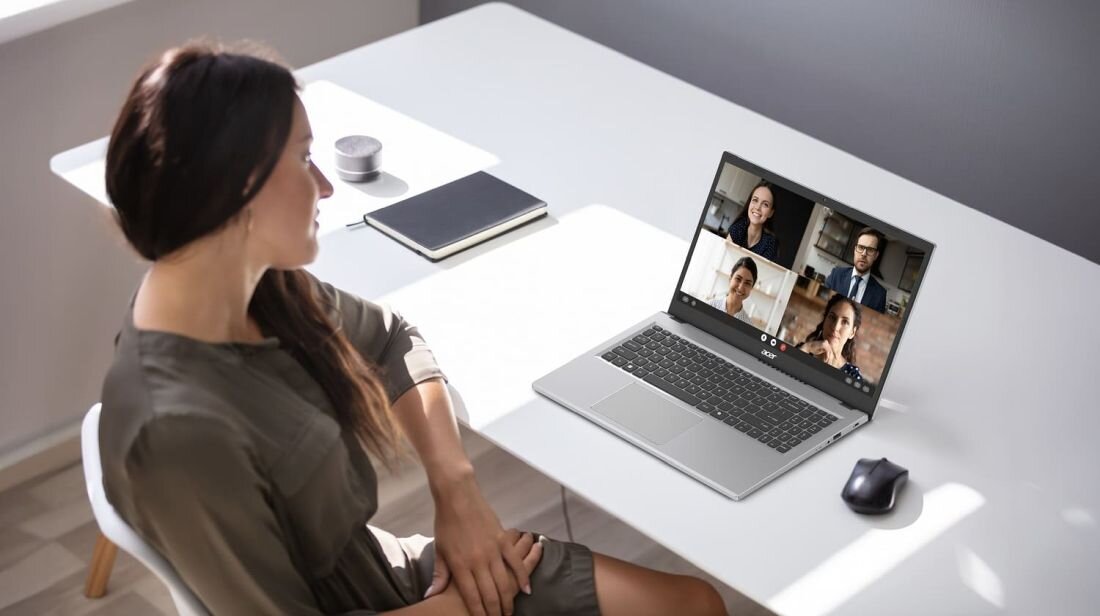 Osoba siedzi przy biurku, na którym znajduje się otwarty laptop z widocznym ekranem, na którym trwa wideokonferencja z czterema osobami. Obok laptopa leży myszka, a na biurku znajdują się również notatnik i filiżanka.