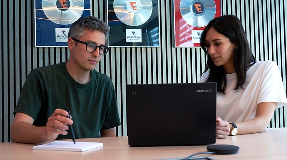 Dwie osoby siedzą przy stole, jedna z nich trzyma długopis, a przed nimi znajduje się laptop marki Acer. W tle widoczne są trzy plakaty z logo \'elasticStage\'.