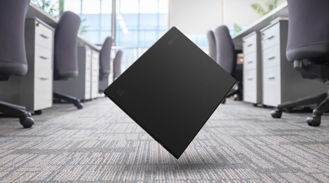 Czarny laptop z logo \'ACER\' na pokrywie, unosi się nad podłogą w biurowym otoczeniu z biurkami i krzesłami.