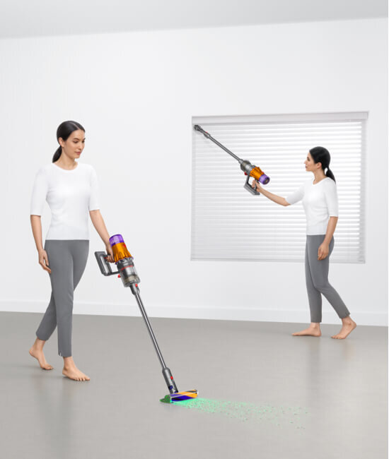 Dwie kobiety używają odkurzacza bezprzewodowego na szarej podłodze i przy oknie z żaluzjami. Odkurzacz ma przezroczysty pojemnik i kolorowe elementy.