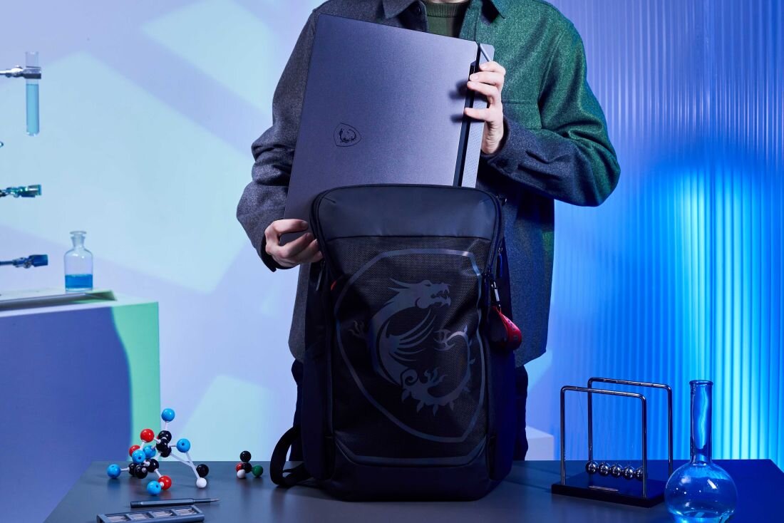 Osoba w szarej marynarce wkłada ciemnoszarego laptopa MSI Vector do czarnego plecaka z logo MSI; w tle nowoczesne laboratorium z modelem cząsteczek i wahadłem Newtona na biurku, co symbolizuje mobilność i zastosowanie w nauce. - Potęga w solidnej obudowie  