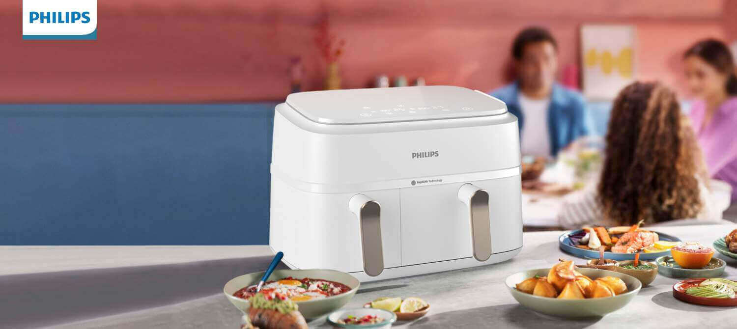 Na pierwszym planie znajduje się biała frytkownica beztłuszczowa z logo PHILIPS, ustawiona na stole z różnymi potrawami. W tle widoczne są rozmyte sylwetki osób siedzących przy stole.