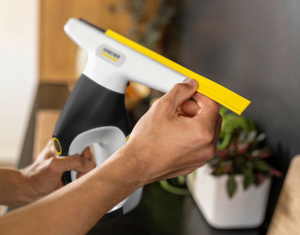 Osoba trzyma myjkę do okien z logo KARCHER, z żółtą gumową końcówką. W tle widoczna jest roślina w białej doniczce.
