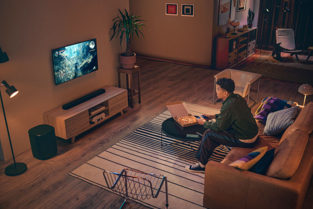 Soundbar JBL BAR 500 MkII wciągający dźwięk kinowy Dolby Atmos przestrzenność, osoba siedząca na kanapie z kontrolerem w ręku, soundbar stojący na komodzie pod zawieszonym telewizorem, subwoofer stojący obok komody
