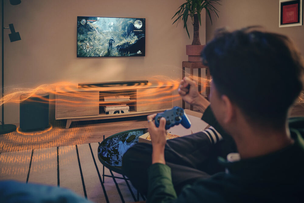 Soundbar JBL BAR 500 MkII 5.1-kanałowy system audio moc 750 W subwoofer 10 cali, osoba siedząca na kanapie z kontrolerem w ręku, soundbar stojący na komodzie pod zawieszonym telewizorem, subwoofer stojący obok komody