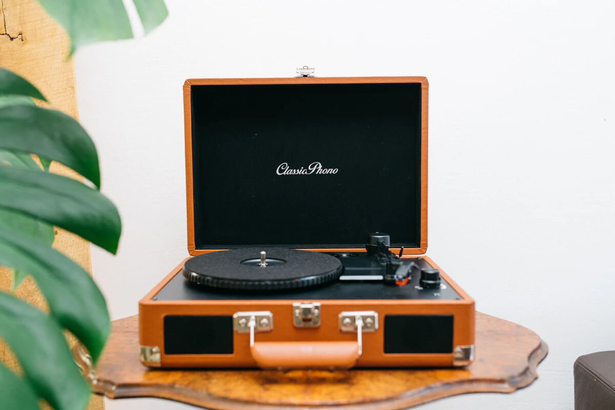 Brązowy gramofon w stylu walizki z otwartą pokrywą, na której widnieje napis \'Classic Phono\'. Gramofon stoi na drewnianym stoliku, a w tle widoczna jest roślina.