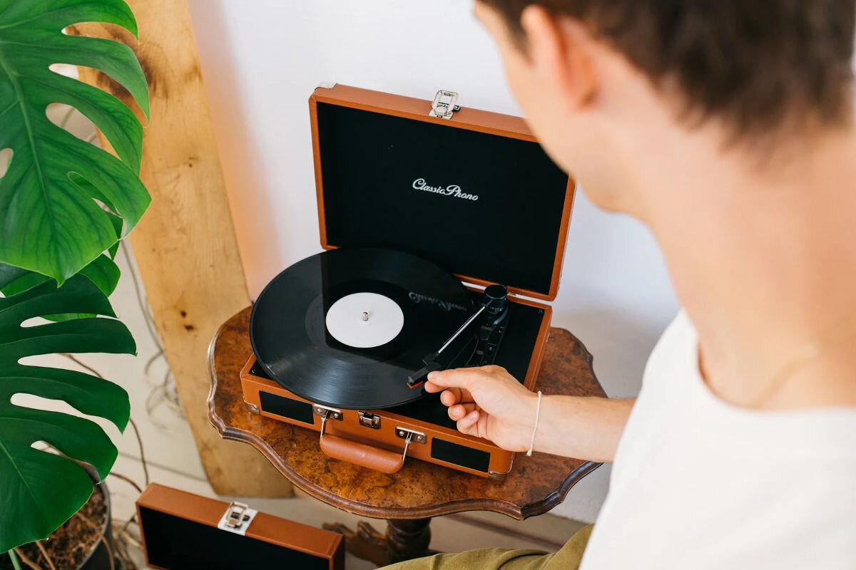 Osoba obsługuje gramofon w brązowej walizce, na którym odtwarzana jest czarna płyta winylowa. Na wewnętrznej stronie pokrywy widoczne jest logo \'Classic Phono\'.