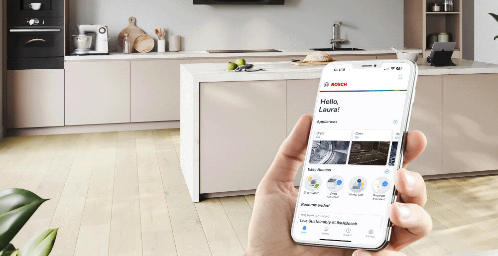 Osoba trzyma smartfon z aplikacją, na której widnieje logo BOSCH i tekst \'Hello, Laura!\'. W tle znajduje się nowoczesna kuchnia z jasnymi szafkami i sprzętem kuchennym.