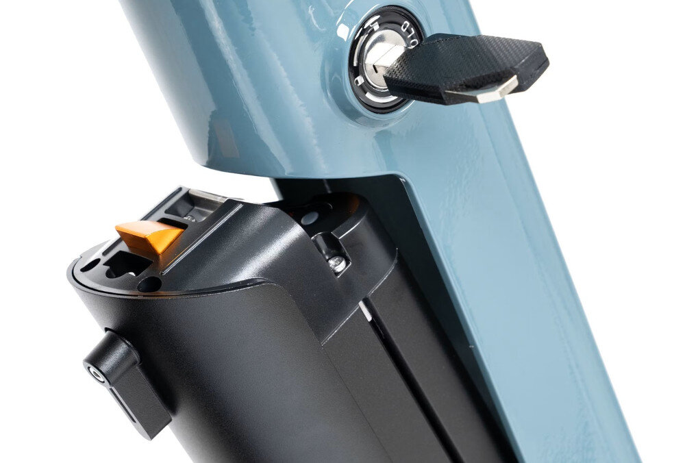 Zbliżenie na zamek z kluczem w rowerze elektrycznym, z widoczną częścią baterii i pomarańczowym przyciskiem. Obudowa jest w kolorze szarym.