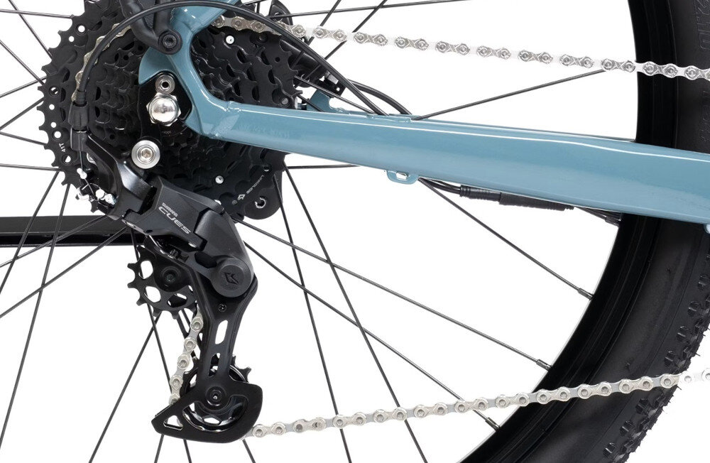 Zdjęcie przedstawia tylną część roweru z widocznym mechanizmem przerzutki i łańcuchem. Rama roweru jest w kolorze szarym, a koło ma czarną oponę.