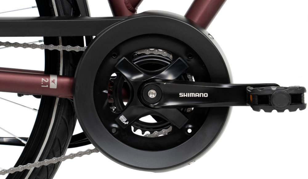 Zbliżenie na mechanizm korbowy roweru z widocznym napisem \'SHIMANO\' na ramieniu korby. Obok znajduje się fragment ramy z oznaczeniem \'2.1\'.