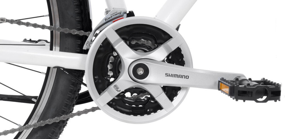 Zbliżenie na mechanizm korbowy roweru z widocznym napisem \'Shimano\' na ramieniu korby. Obok znajduje się pedał z odblaskiem oraz fragment łańcucha i opony.