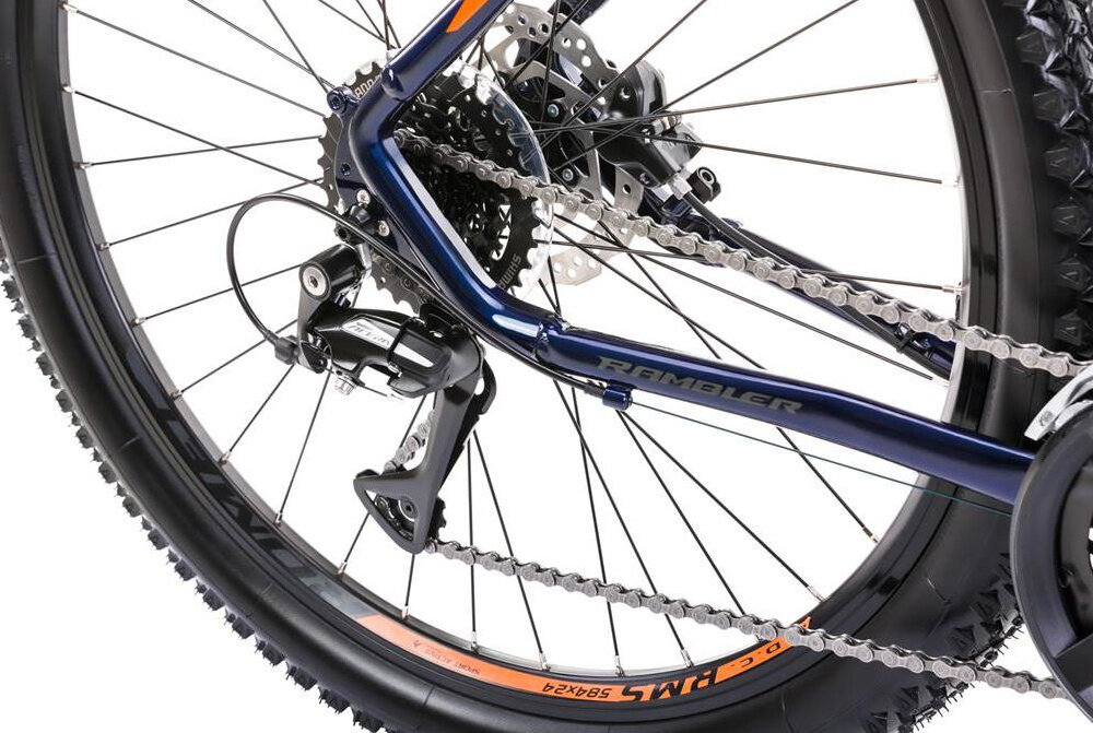 Zbliżenie na tylne koło roweru górskiego z widocznym mechanizmem przerzutki i łańcuchem. Na ramie widoczny jest napis \'RAMBLER\'.