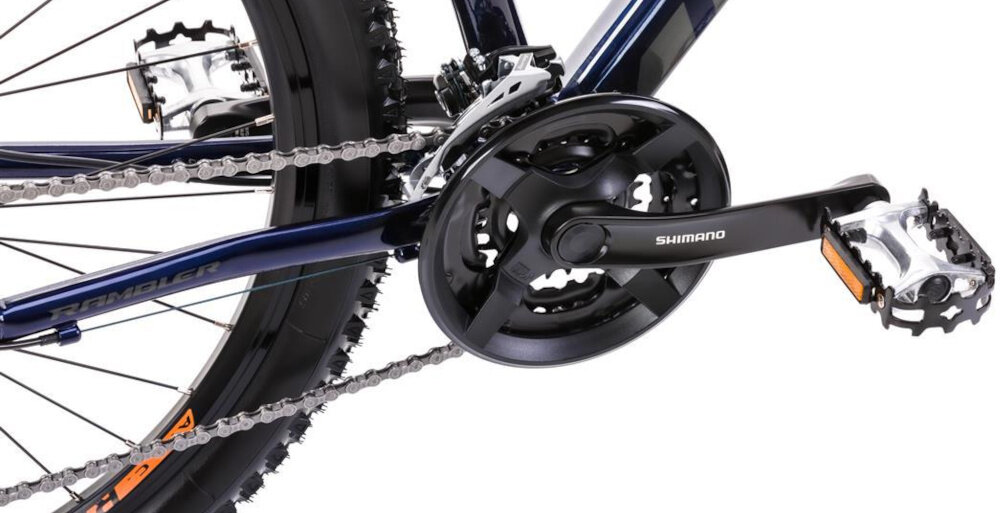 Zbliżenie na mechanizm napędowy roweru górskiego z widocznym napisem \'SHIMANO\' na korbie. Rama roweru jest granatowa z pomarańczowymi akcentami i napisem \'RAMBLER\'.