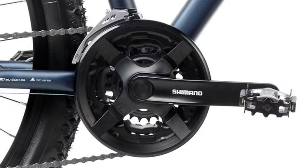 Zbliżenie na mechanizm korbowy roweru z widocznym napisem \'SHIMANO\' na ramieniu korby. Łańcuch jest zamontowany na zębatce, a rama roweru ma granatowy kolor.