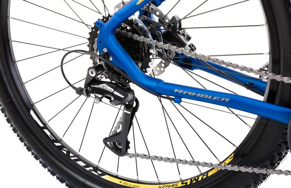 Zbliżenie na tylne koło roweru górskiego z niebieską ramą i widocznym napisem \'RAMBLER\'. Widać mechanizm przerzutki oraz opony z żółtymi detalami.