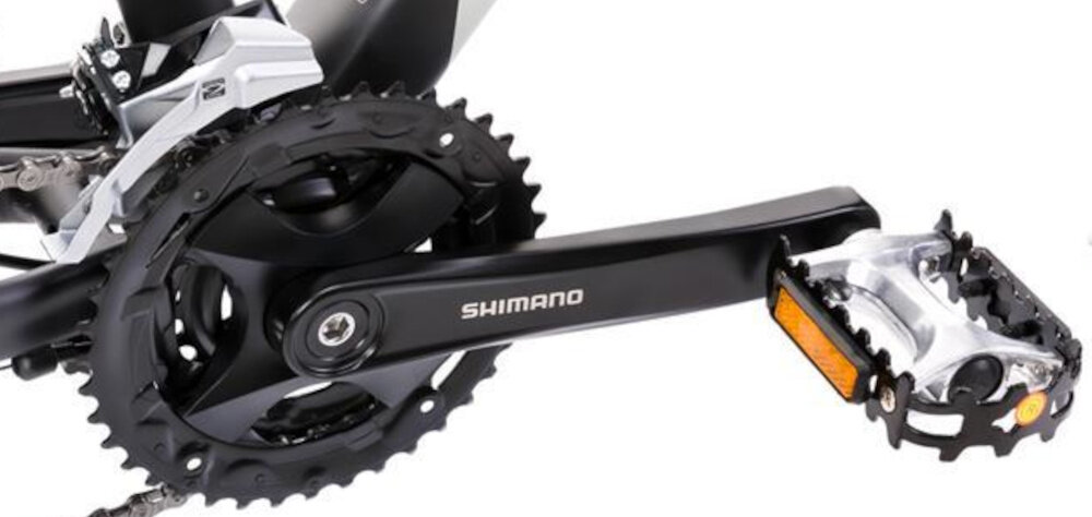 Zdjęcie przedstawia mechanizm korbowy roweru z widocznym napisem \'SHIMANO\' na ramieniu korby. Obok znajduje się pedał z elementem odblaskowym.